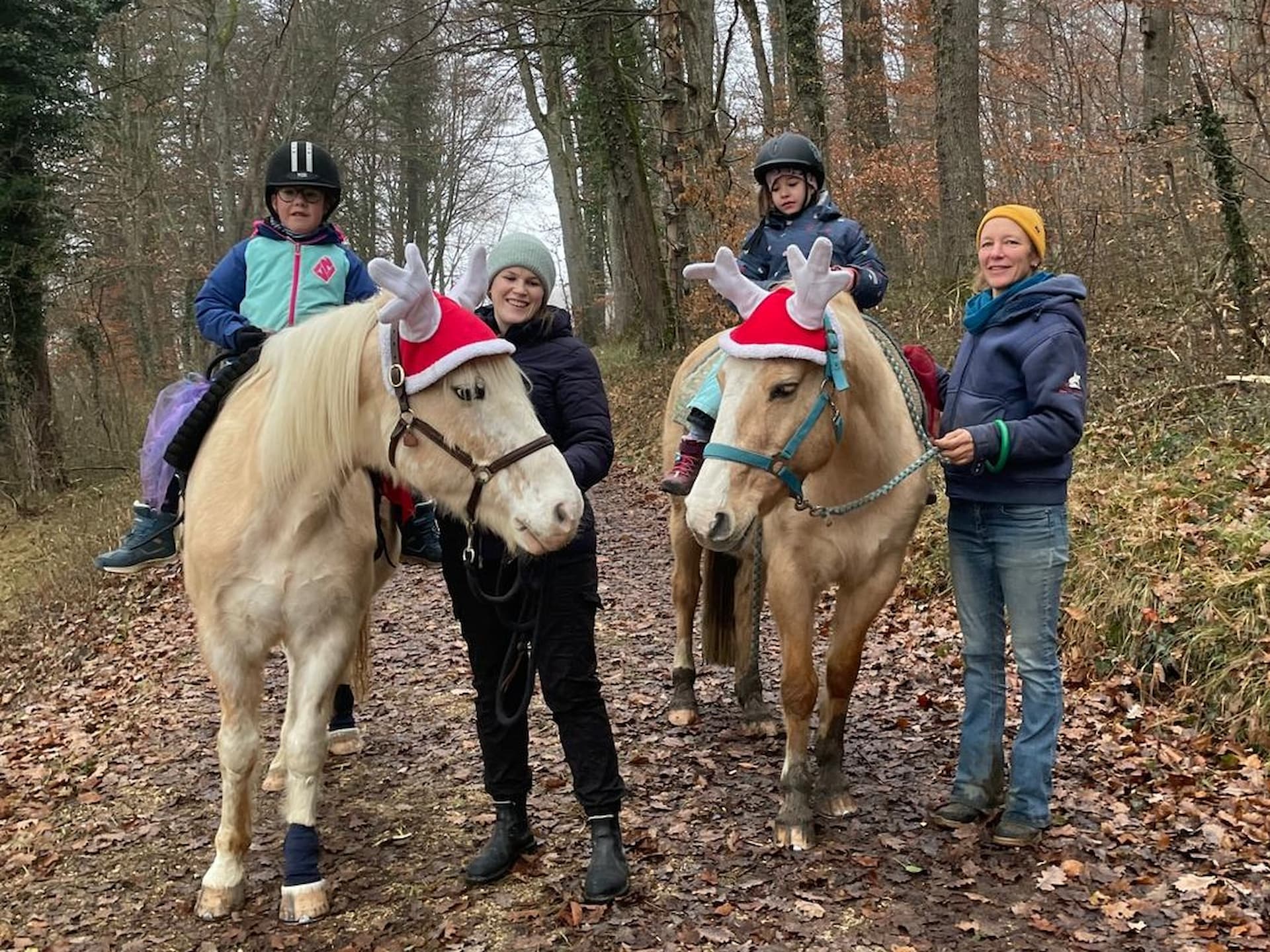 Zwei Kinder reiten festlich geschmückte Pferde im Wald. Zwei Frauen führen dabei die Pferde.